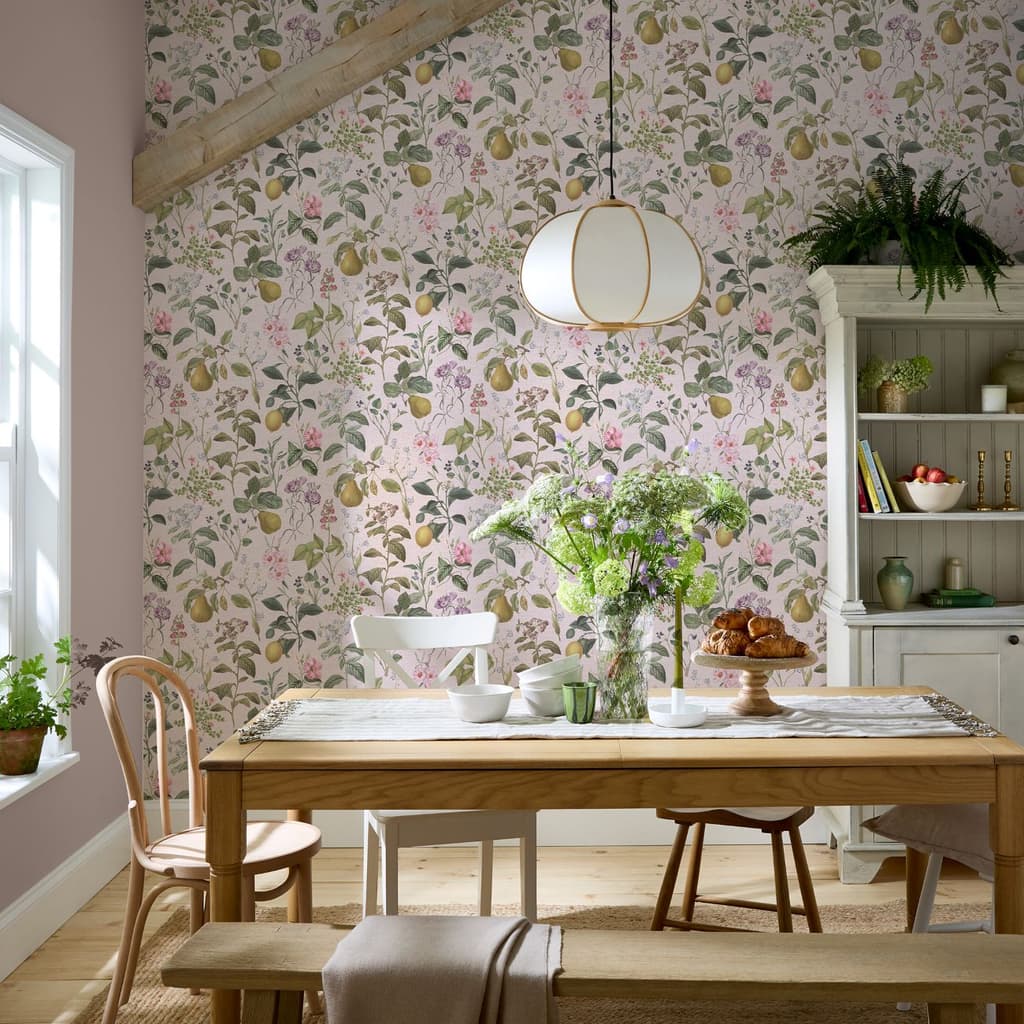 Superfresco Easy Ophelia Pink Wallpaper in a dining room with wooden dining table and pale wood tall cupboard with open shelves.